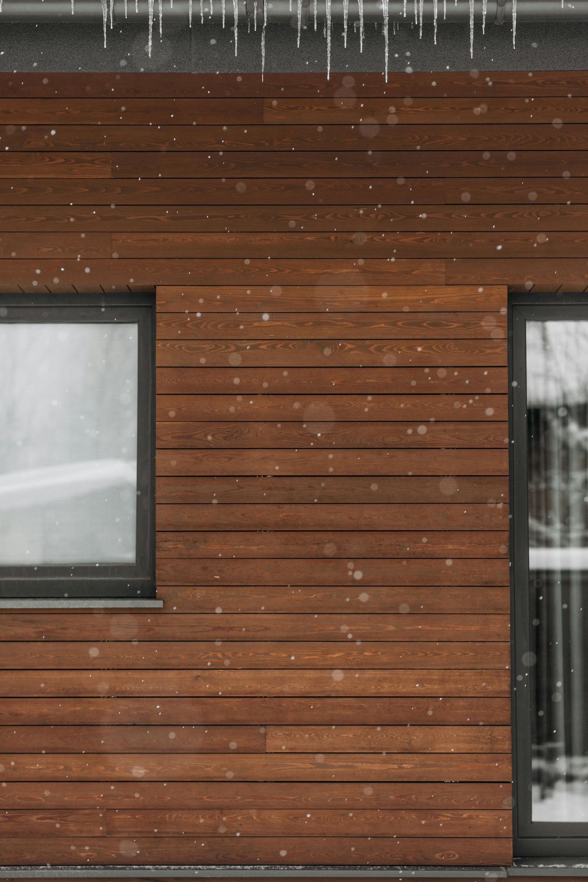 La neige tombe à l'extérieur d'une maison moderne avec un revêtement extérieur en bois brun et des fenêtres