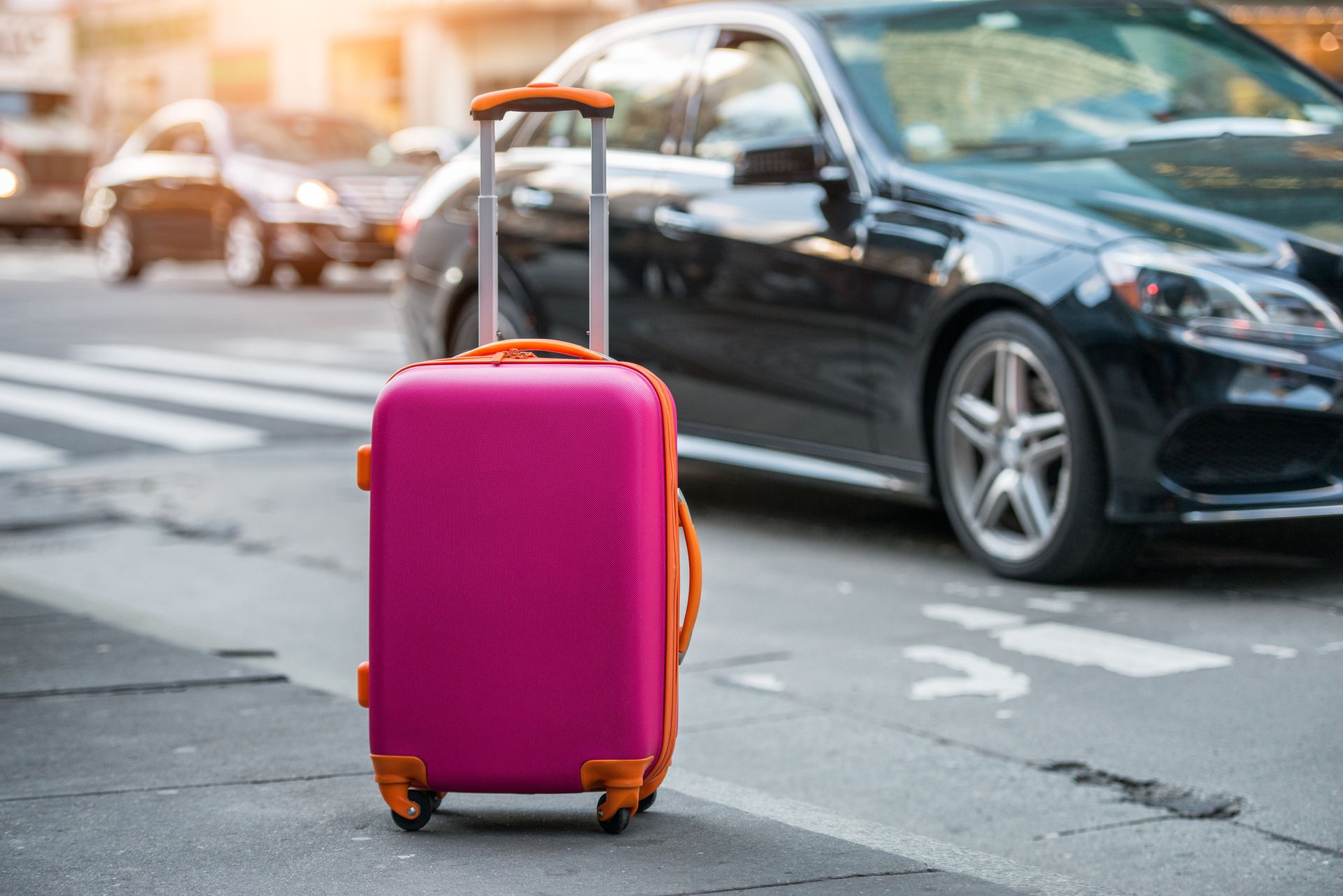 Ein rosa Koffer steht auf einer Strasse in der Stadt neben geparkten Autos.