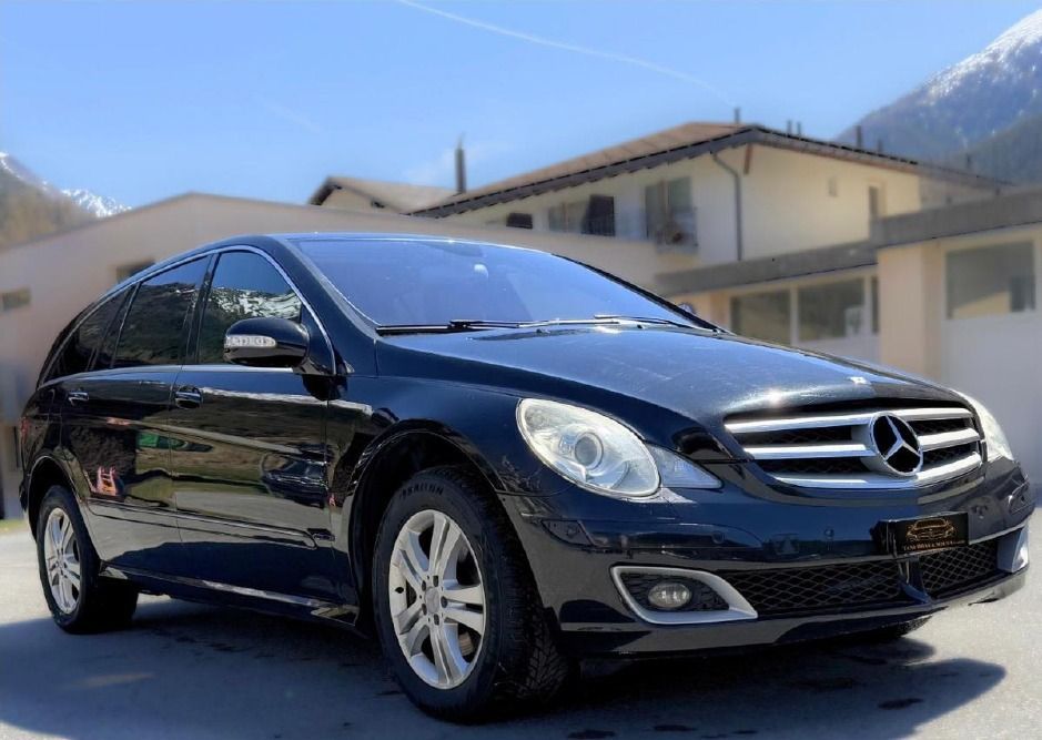 Eine schwarze Mercedes-Benz Limousine parkt in einer sonnigen Strasse; im Hintergrund sind Berge und ein Haus zu sehen.