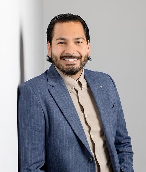 Mann im blauen Blazer lächelt, an einer weißen Wand lehnend. Er hat dunkles Haar und einen Bart.