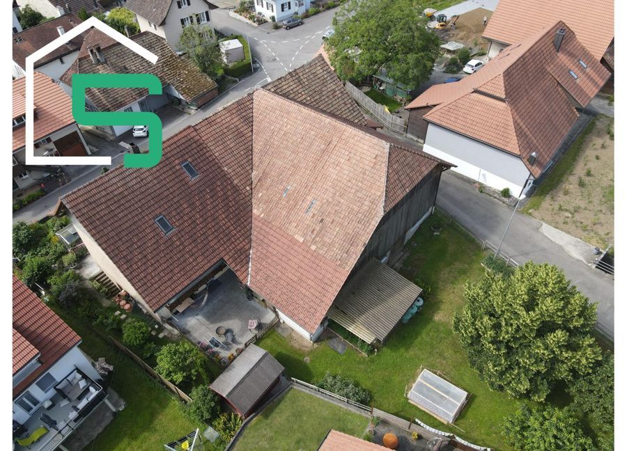 Luftaufnahme eines Gebäudes mit braunem Dach in einem Dorf und einem grünen Logo in der oberen linken Ecke.