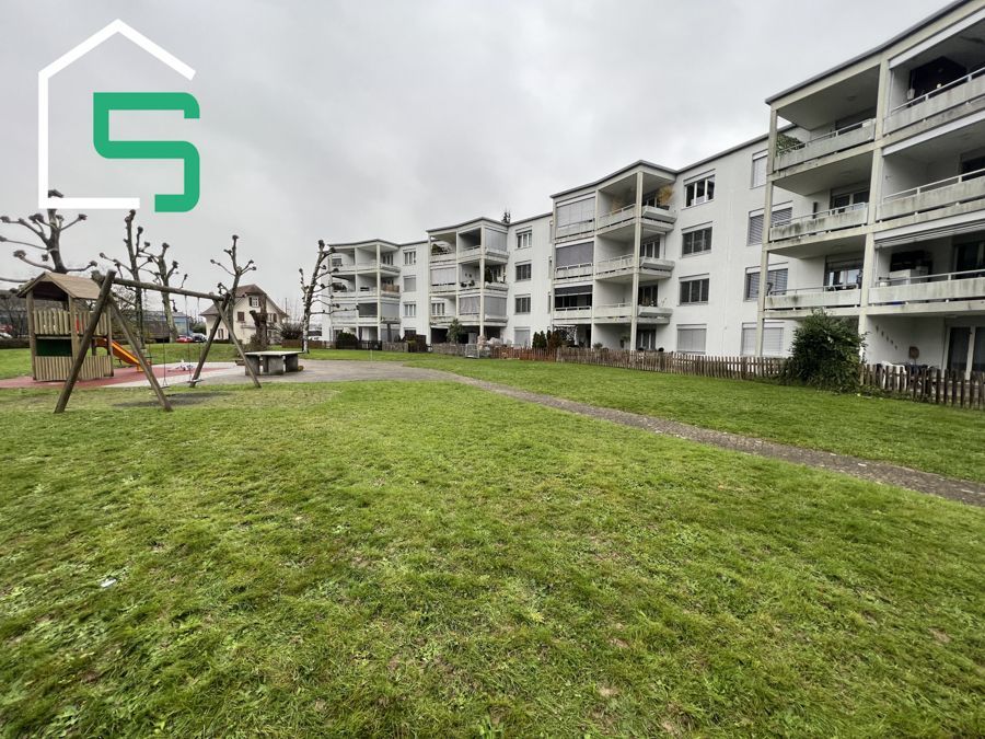Apartmentkomplex mit Balkonen und einem Spielplatz auf einer Rasenfläche. Bewölkter Himmel.