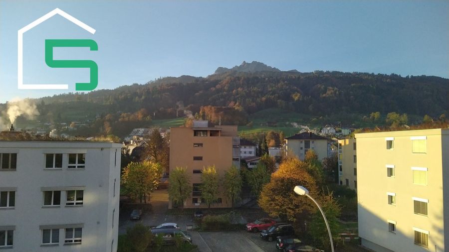 Gebäude in einer Stadt mit einem Berg im Hintergrund unter einem sonnigen, klaren Himmel. Logo in der oberen linken Ecke.