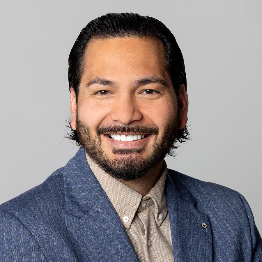 Mann mit dunklem Haar und Bart, lächelnd, in blauem Nadelstreifenanzug und hellbraunem Button-Down-Hemd.