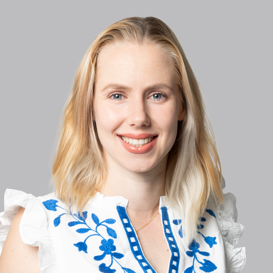 Frau mit blondem Haar, die ein weißes Oberteil mit blauer Stickerei trägt, lächelt vor einem grauen Hintergrund.