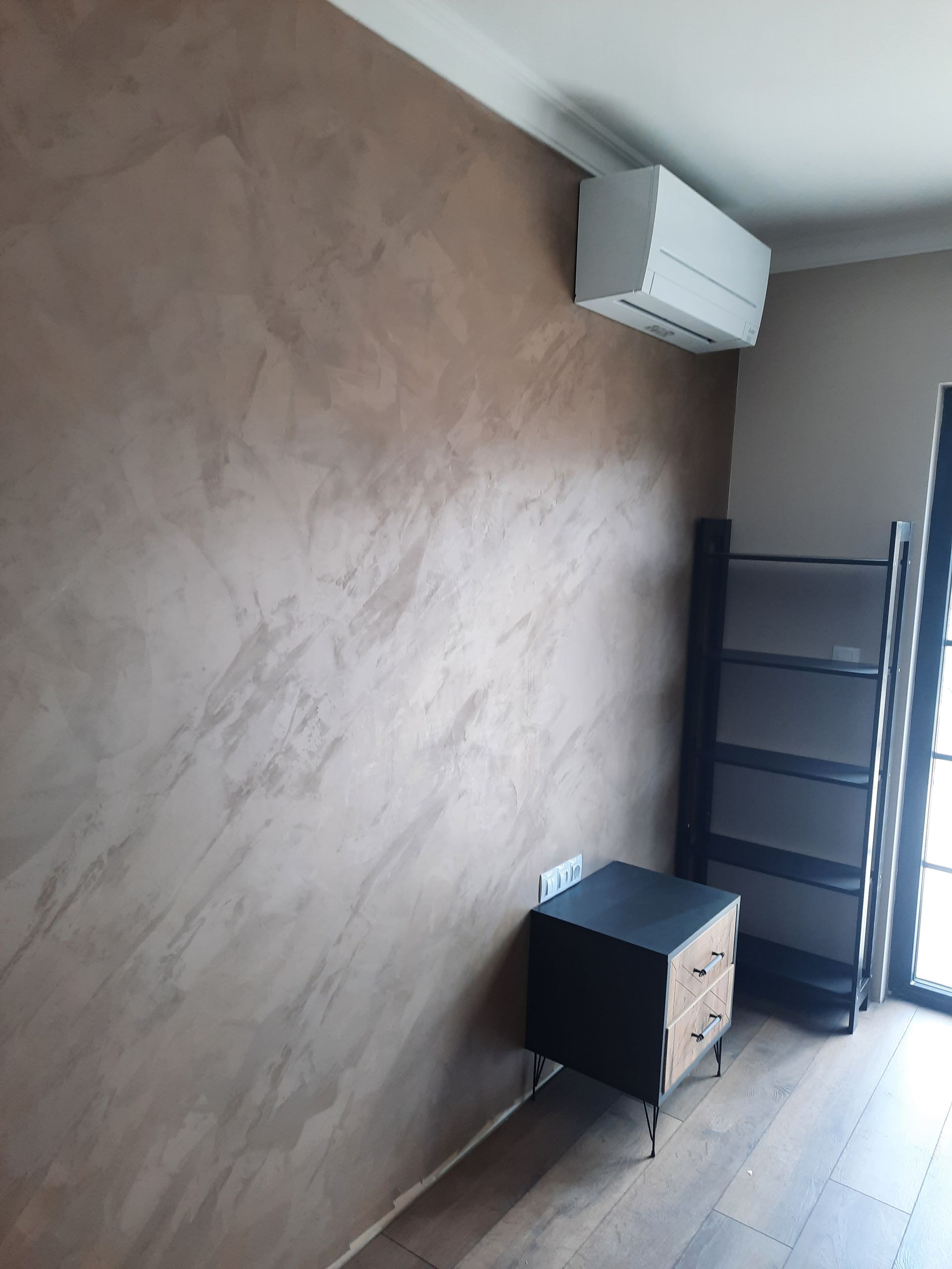 Habitación con pared texturizada marrón, aire acondicionado, mesita de noche, estante y puerta.