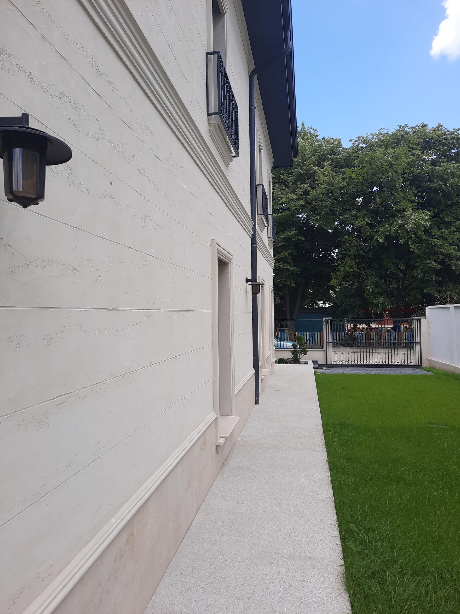 Vista lateral de un edificio de color beige claro con una pasarela, césped verde y árboles bajo un cielo azul.