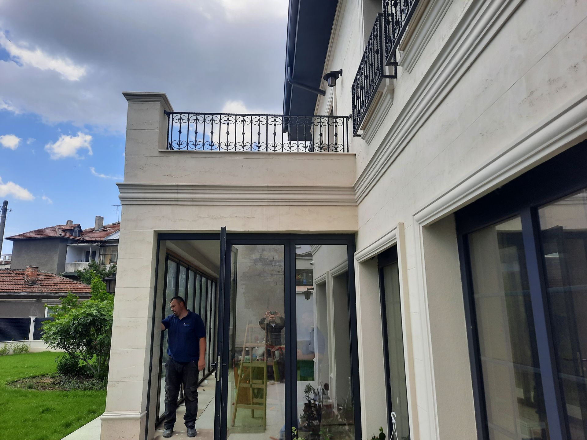 Hombre de pie cerca de las puertas de cristal abiertas de una casa de color claro con detalles en negro y un pequeño balcón.