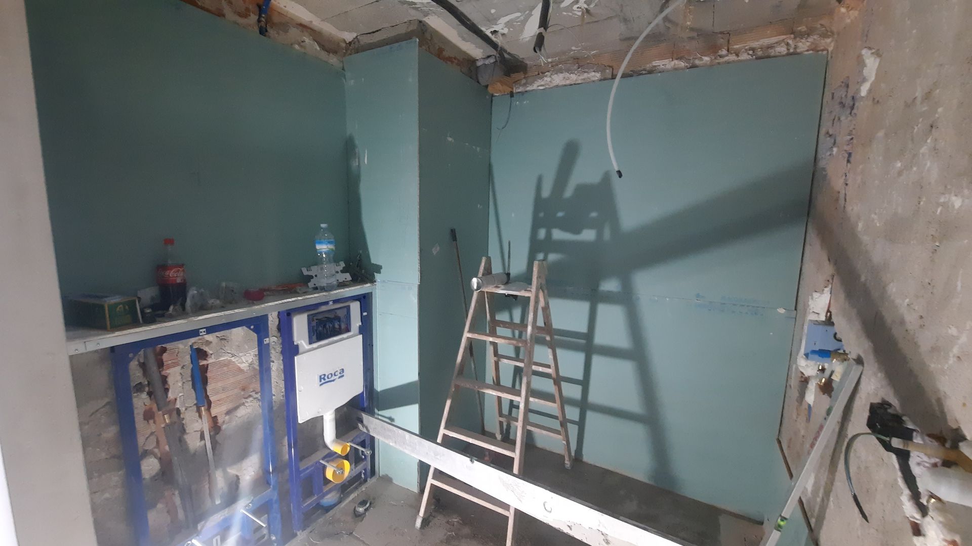Baño en construcción, paredes de pladur azul, instalación de fontanería para el inodoro, escalera presente.