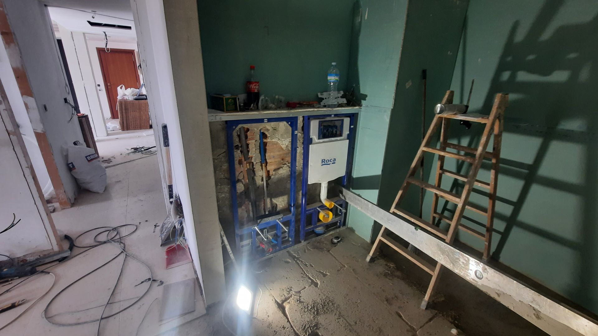 Baño en construcción: tuberías y accesorios a la vista, escalera y herramientas. Paredes verdes, suelo sin terminar.