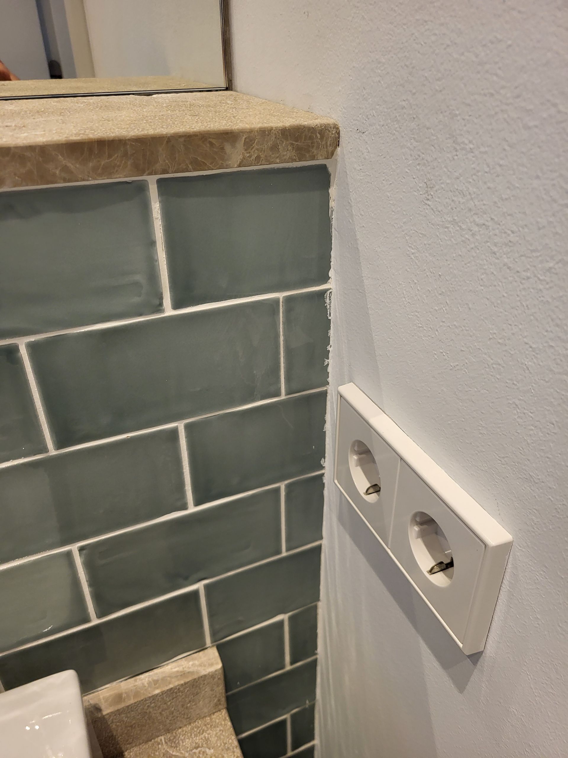 Enchufe doble en pared blanca. Pared alicatada en verde con molduras beige.