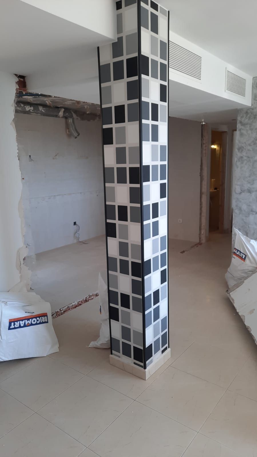 Renovación en curso: Columna revestida de azulejos blancos y negros, habitación con pared a la vista, bolsas en el suelo.