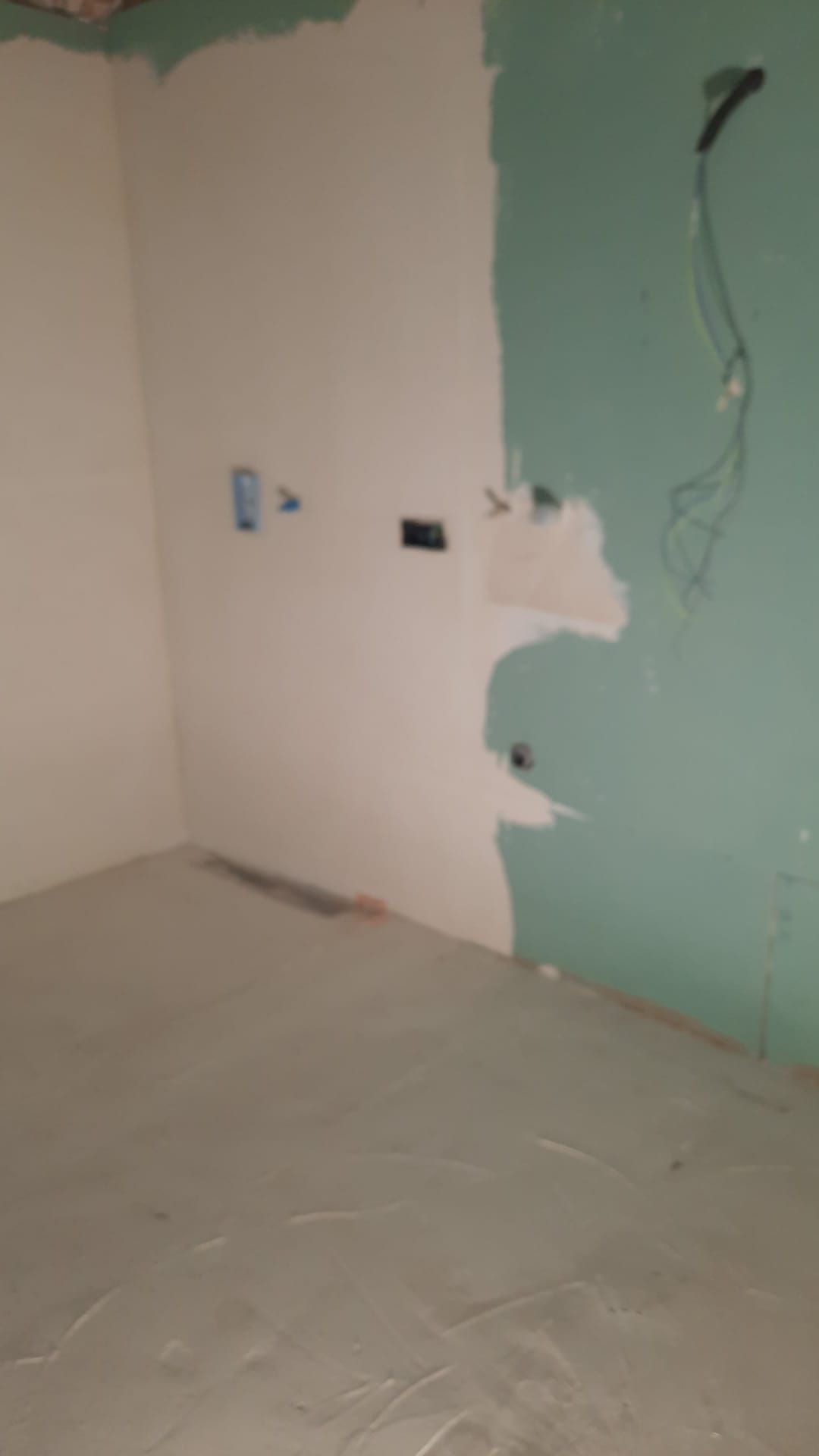 Habitación con paredes sin terminar: paneles de yeso blancos y verdes, enchufes eléctricos y suelo gris.