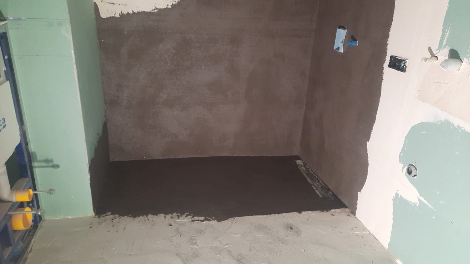 Cabina de ducha en construcción; paredes y suelo de cemento gris, tuberías a la vista.