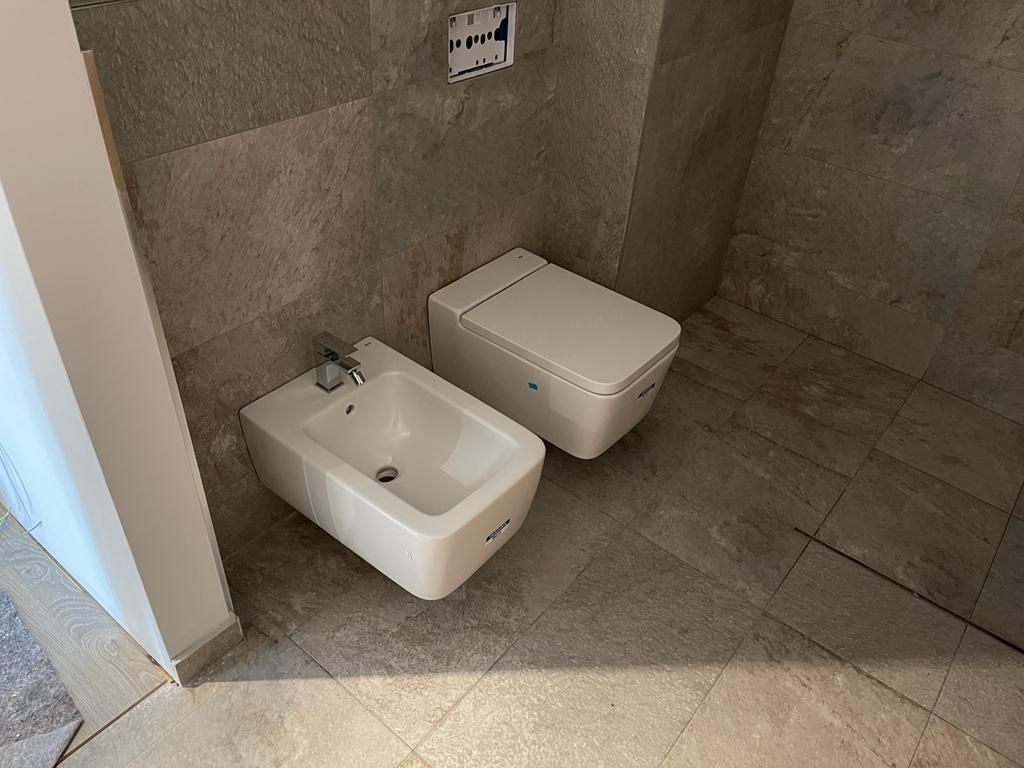 Baño moderno con bidé e inodoro, ambos blancos, sobre suelo de baldosas grises.