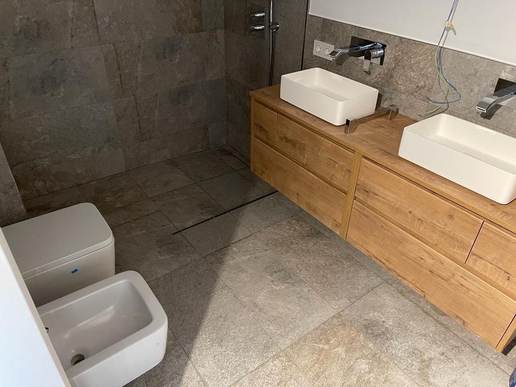 Baño con tocador de madera, dos lavabos, inodoro y ducha alicatada.