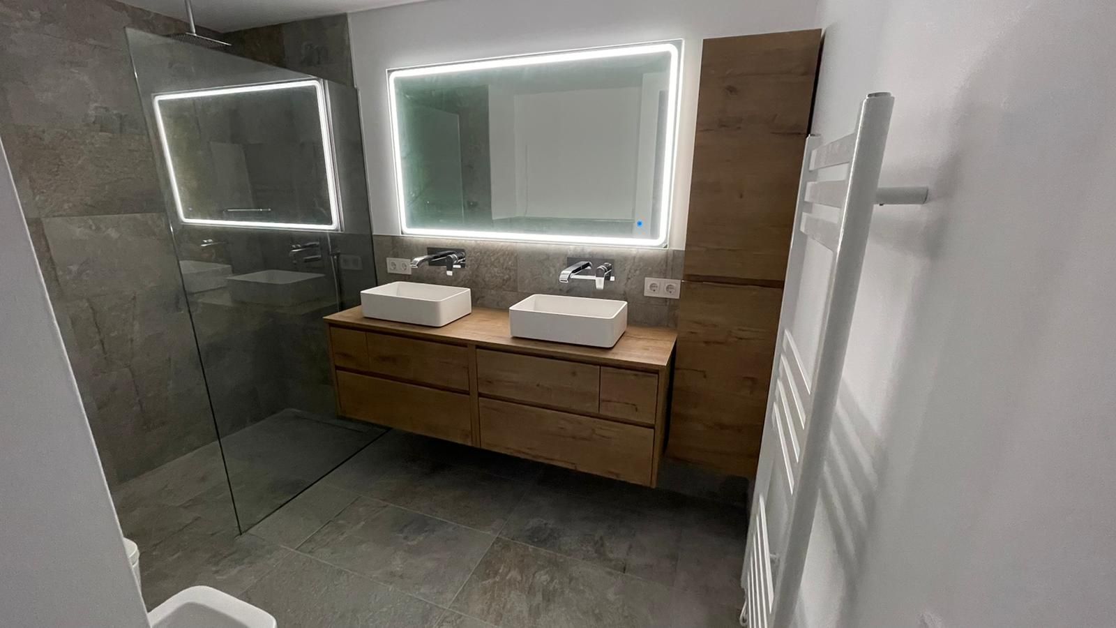 Baño moderno con tocador de madera, dos lavabos blancos, espejo grande y ducha de cristal.