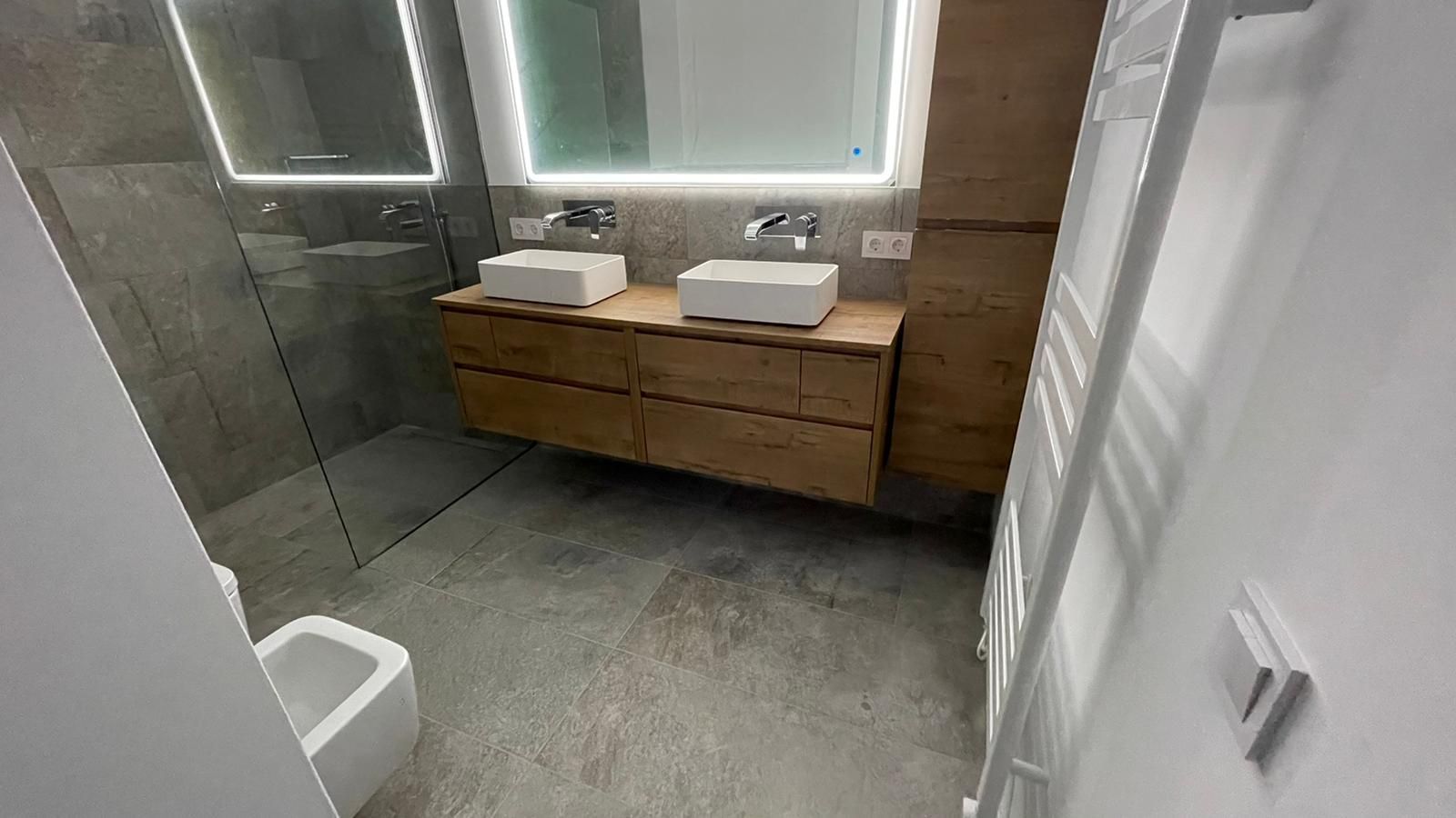 Baño moderno con paredes grises, tocador de madera, dos lavabos y mampara de ducha de cristal.