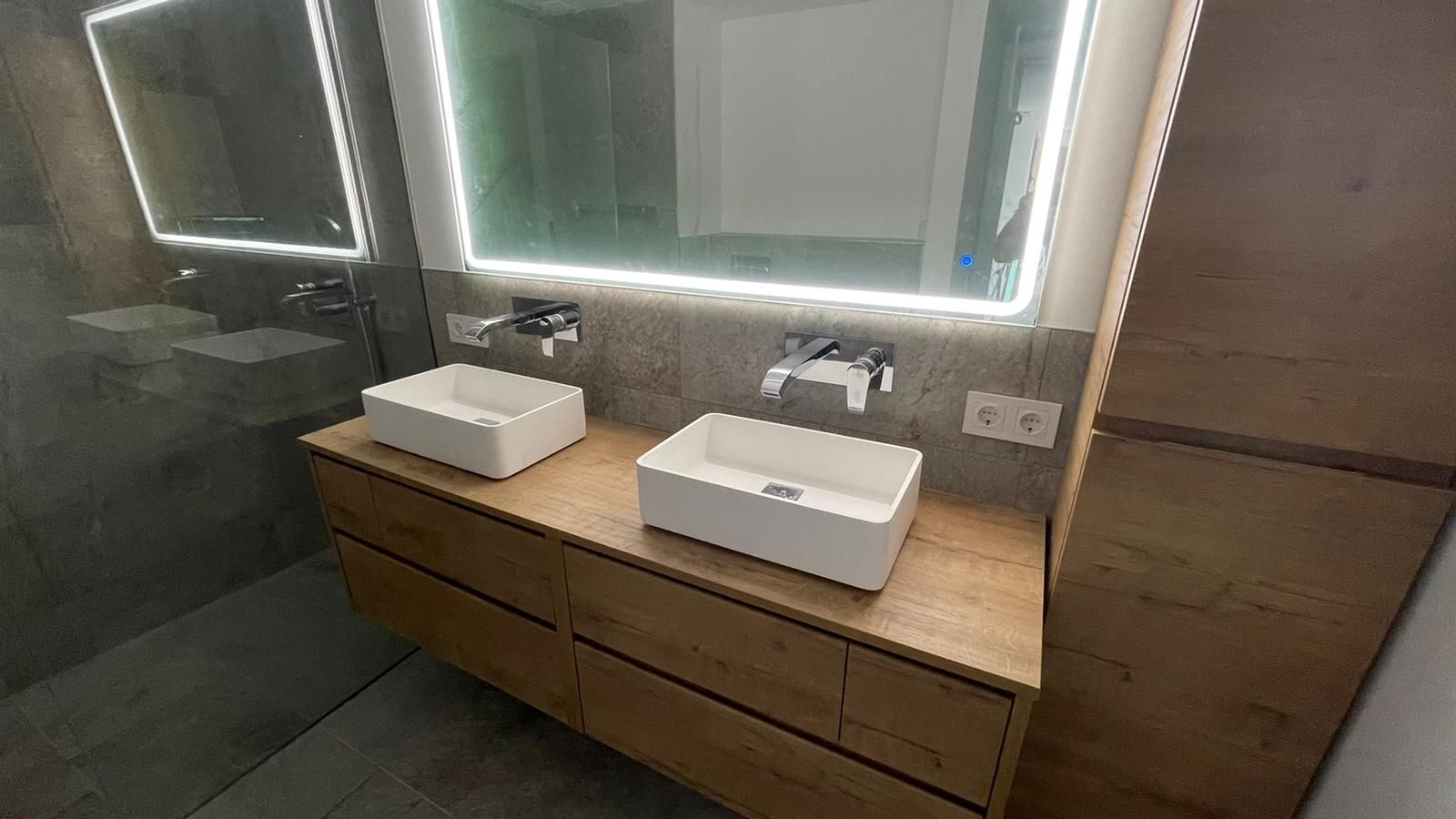 Mueble de baño con dos lavabos blancos, armario de madera y espejo iluminado.
