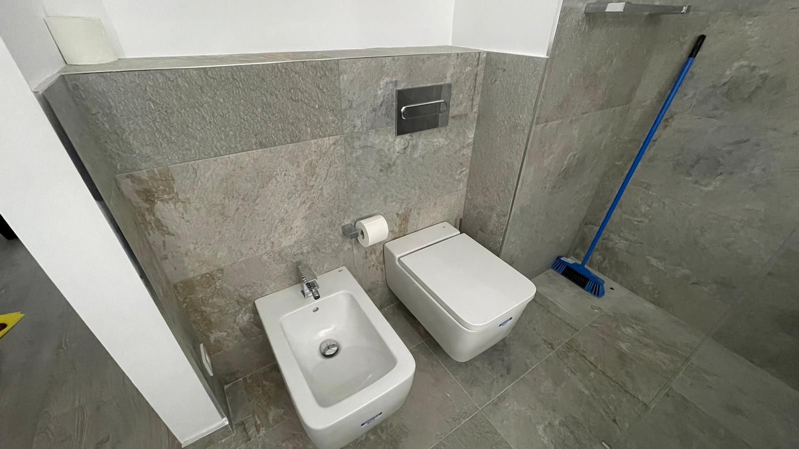 Baño con paredes y suelo de azulejos grises; incluye inodoro, bidé y escoba azul.