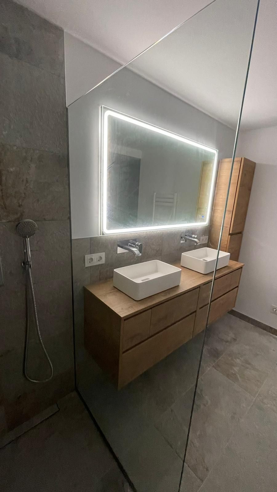 Baño moderno con tocador de madera, lavabos rectangulares, espejo retroiluminado y mampara de ducha de cristal.