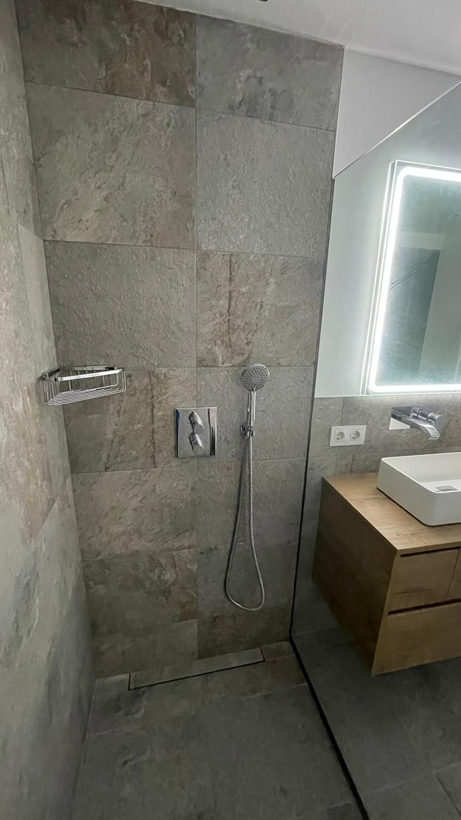 Zona de ducha con paredes de azulejos de piedra, grifería metálica y mampara de cristal.