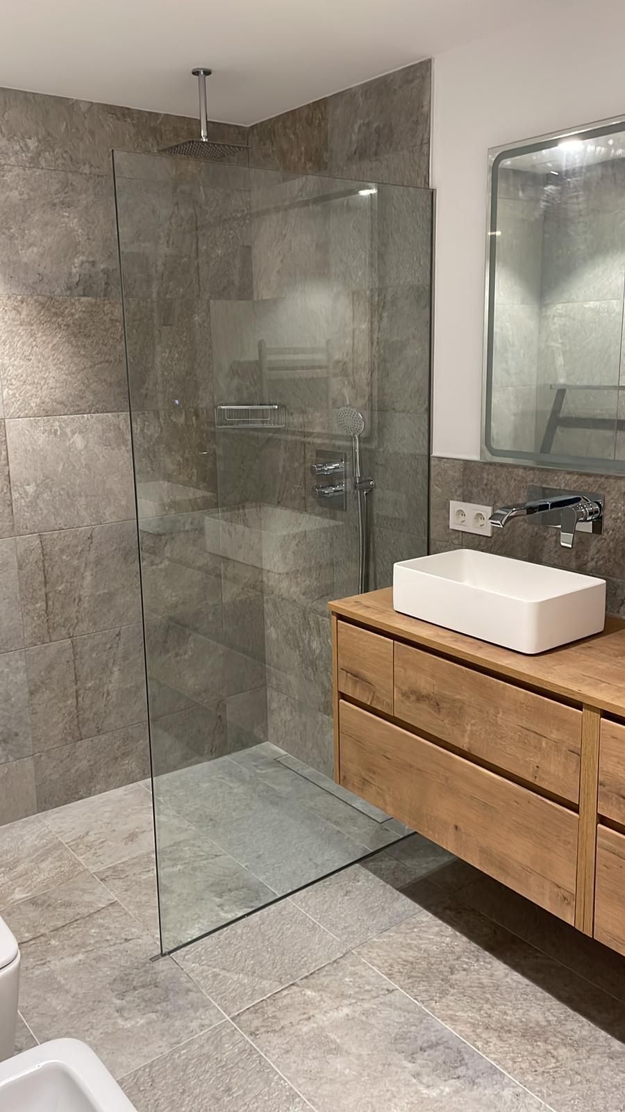 Baño moderno con tocador de madera, lavabo rectangular, ducha de cristal y paredes de piedra.