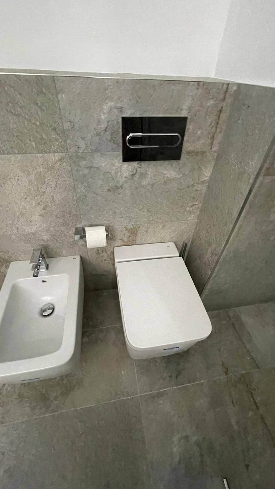 Baño con bidé, inodoro moderno y portarrollos de papel higiénico en una pared de azulejos grises.