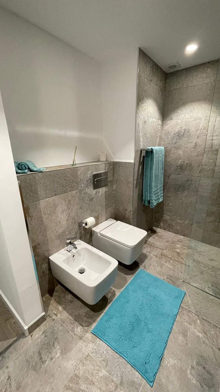 Baño moderno con bidé, inodoro y alfombra azul. Se aprecian las paredes alicatadas en gris y una toalla color verde azulado.