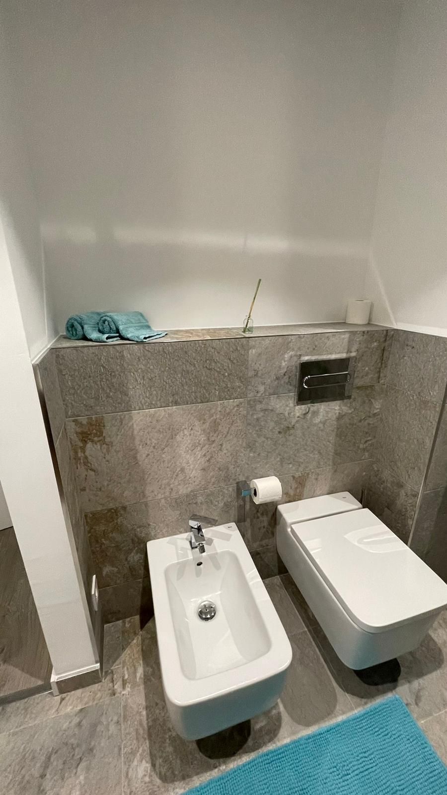 Baño con bidé e inodoro blancos contra una pared de piedra gris, alfombra azul en el suelo.