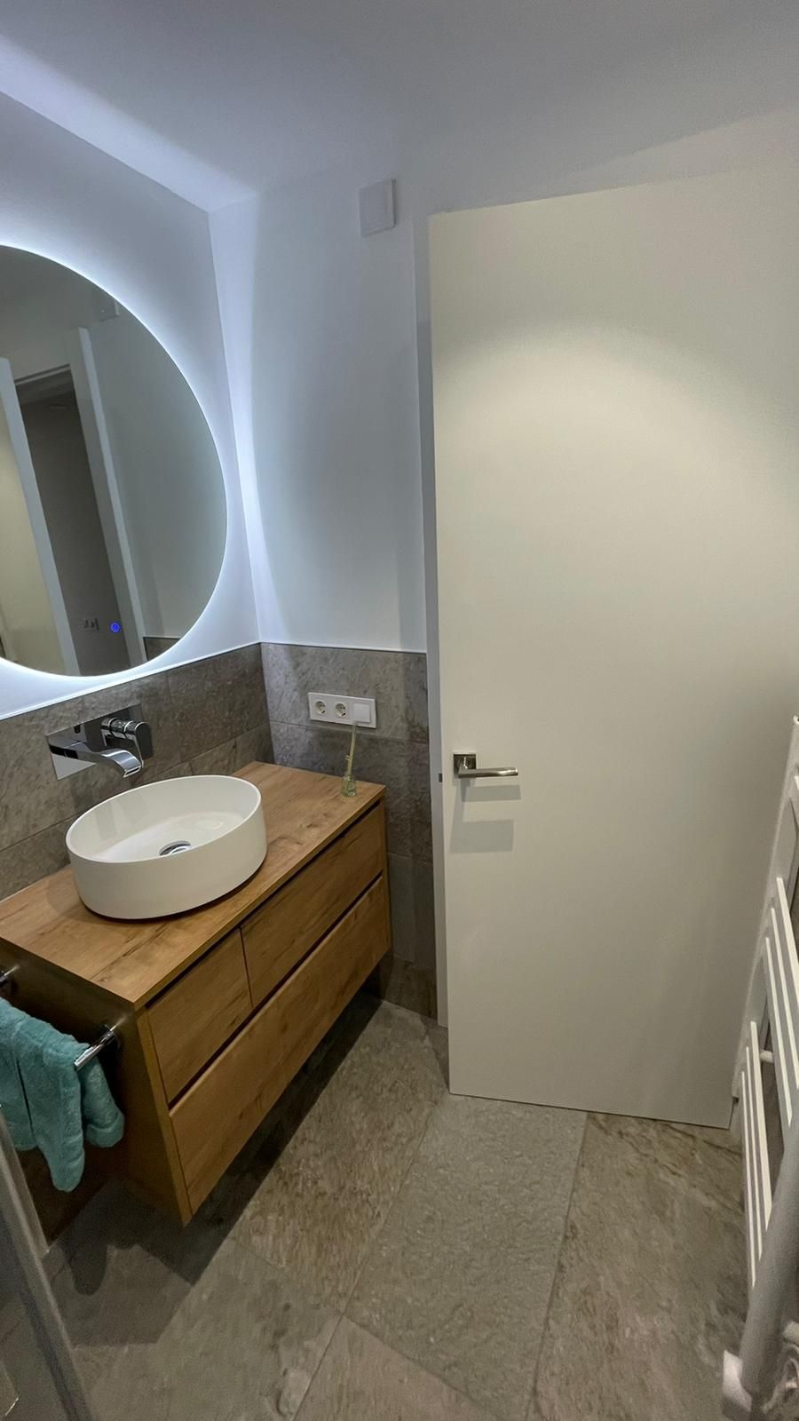 Baño con espejo redondo, tocador de madera con lavabo blanco y puerta blanca.