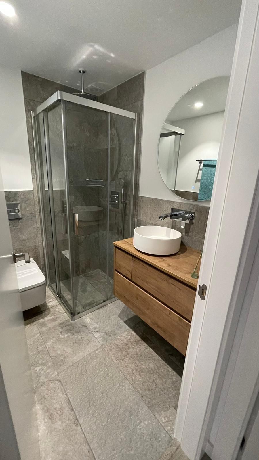 Baño moderno con ducha, lavabo, inodoro, espejo redondo y armario de madera.