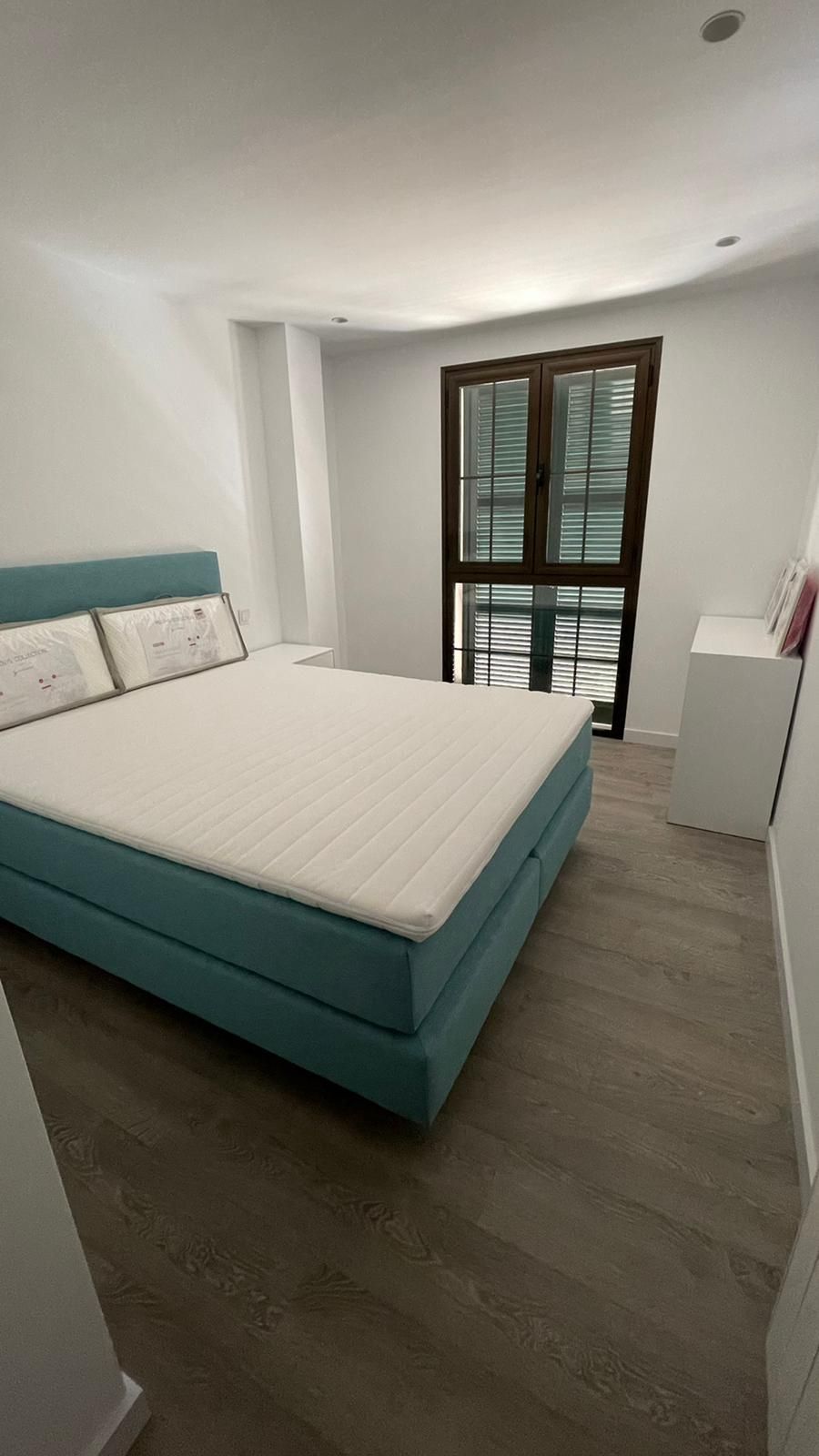 Dormitorio con cama turquesa, colchón blanco y ventana oscura. Suelo de madera clara, paredes blancas.