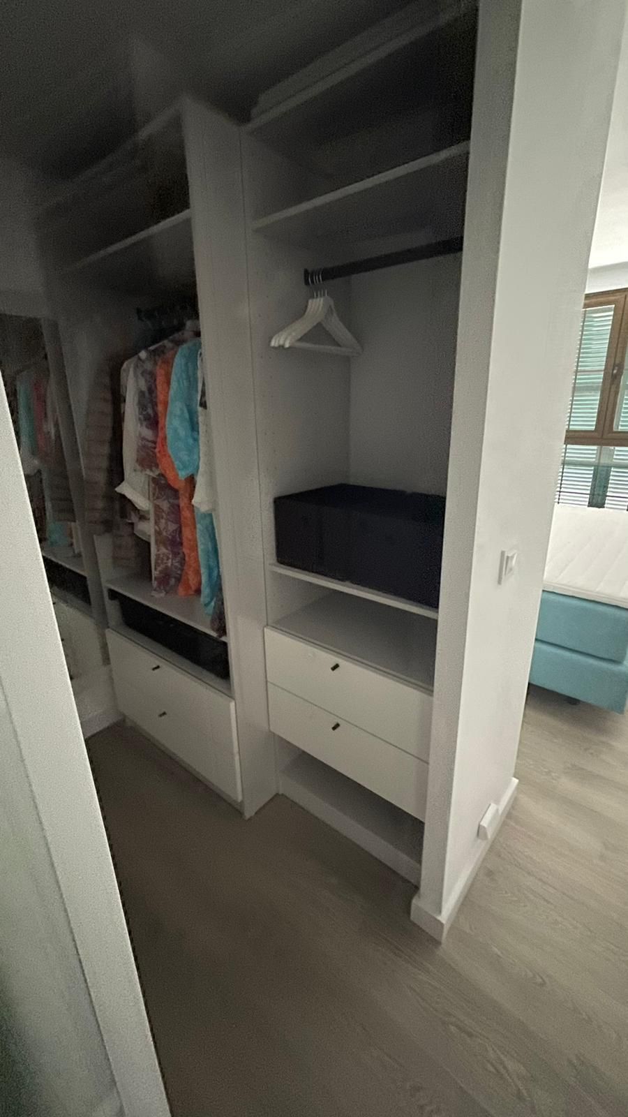 Armario blanco con cajones, estantes y barra para colgar ropa. La ropa queda a la vista. Suelo de madera clara.