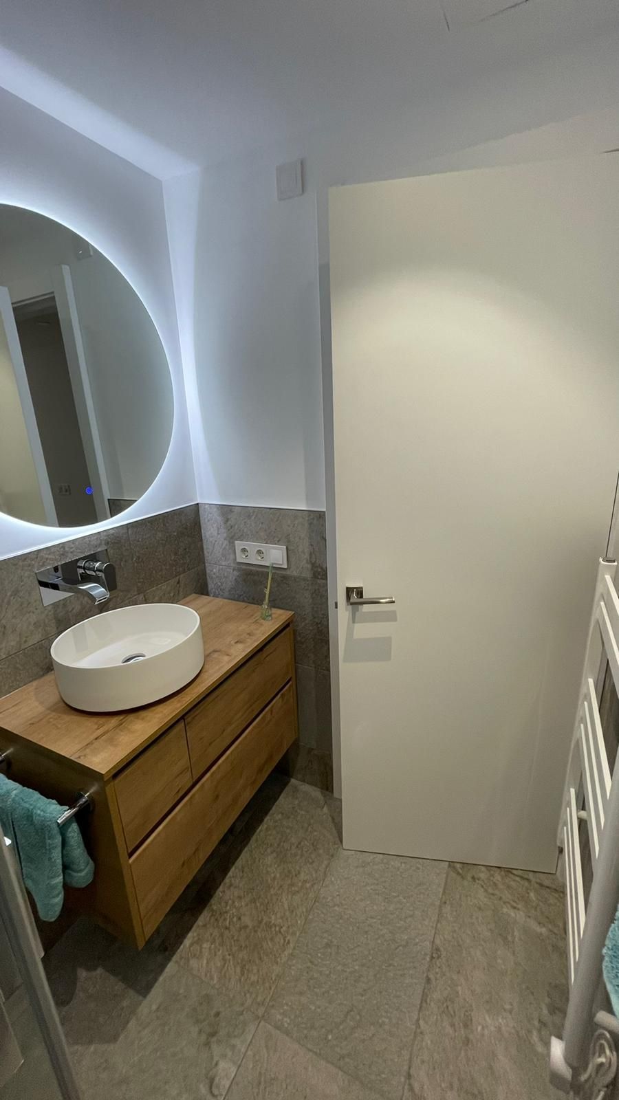 Baño con tocador de madera, lavabo redondo, espejo circular con LED y puerta blanca.
