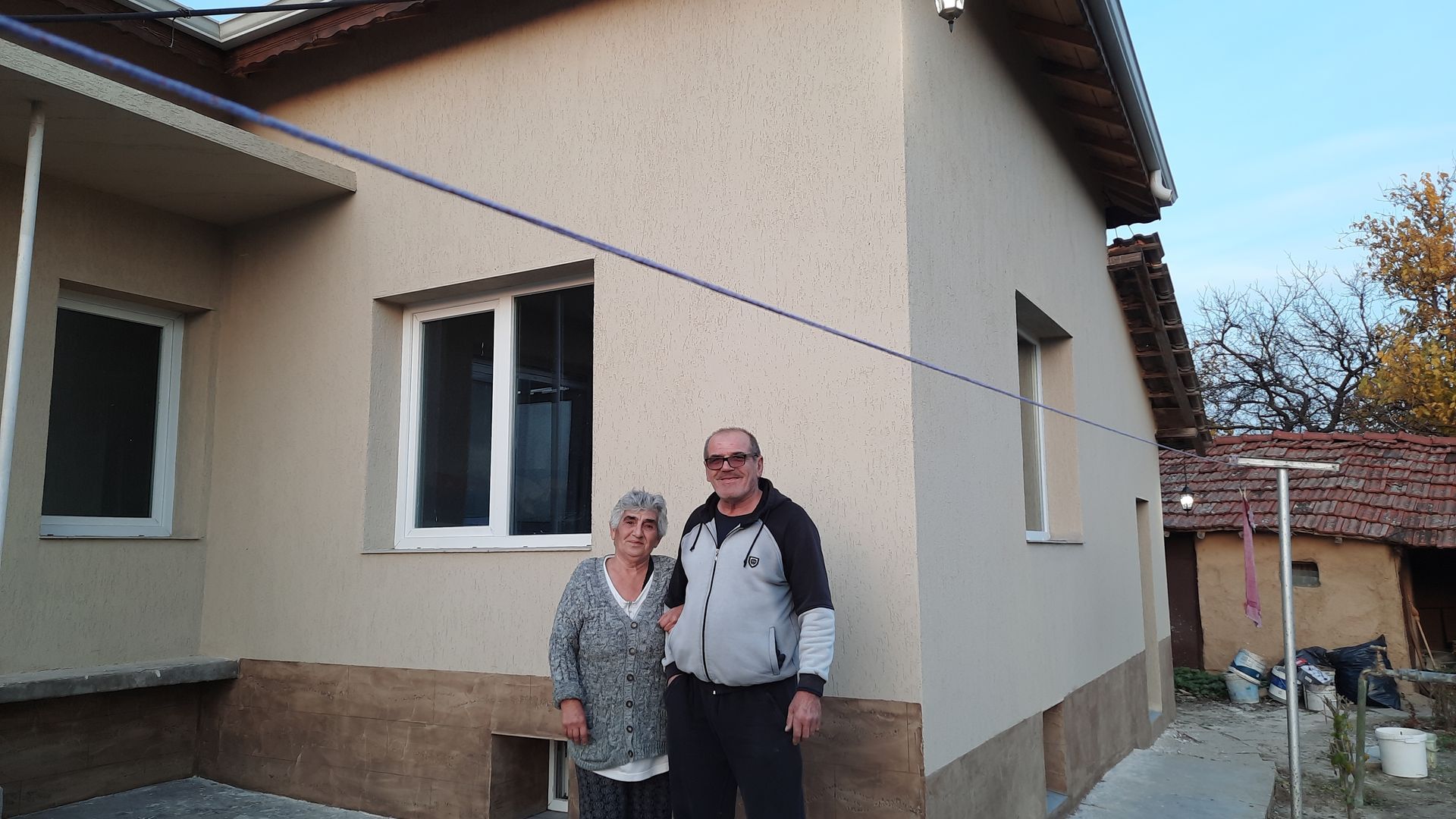 Pareja de pie frente a una casa color beige con marcos de ventanas blancos; cielo azul al fondo.