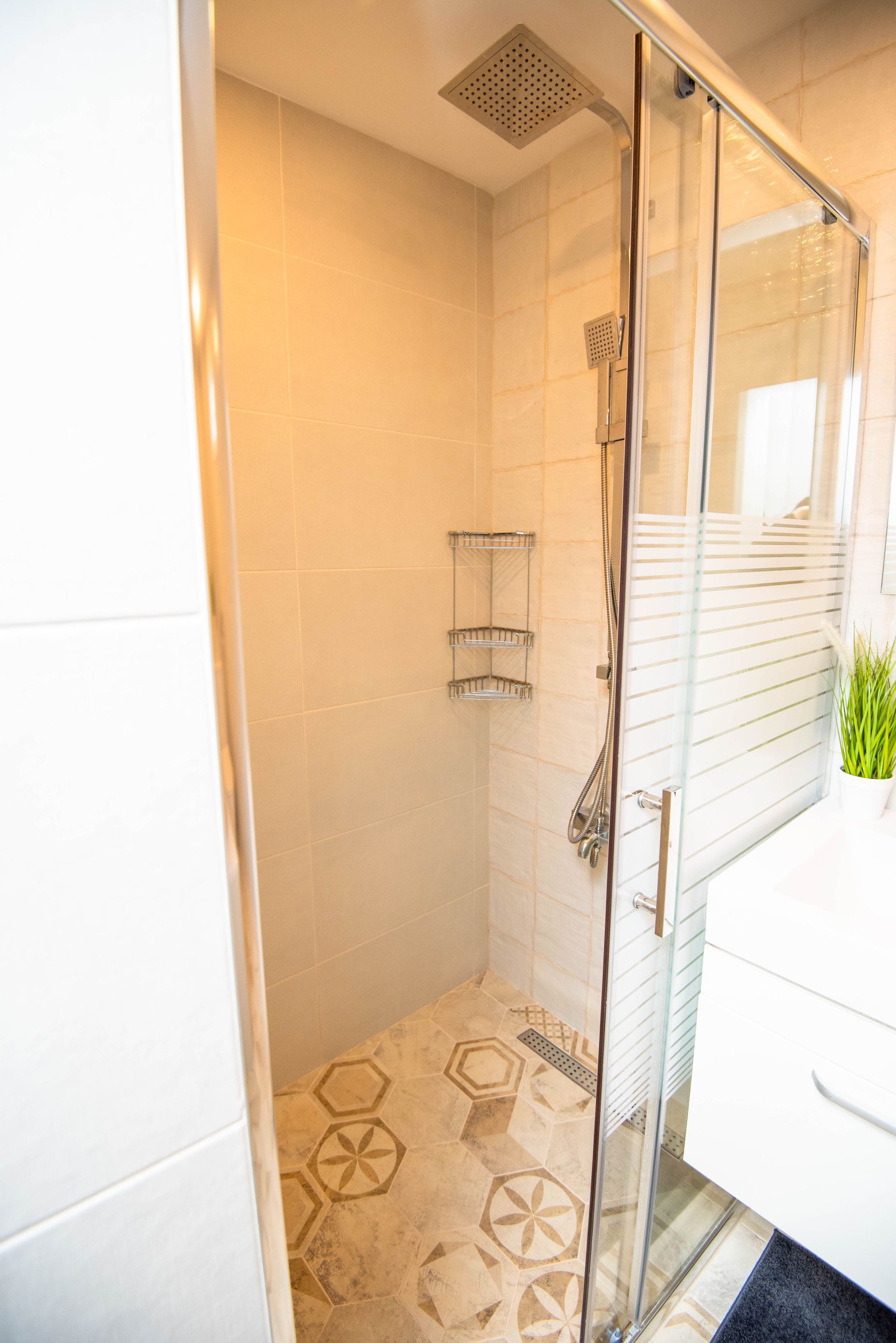 Cabina de ducha con puerta de cristal, paredes alicatadas en blanco y suelo de baldosas estampadas.