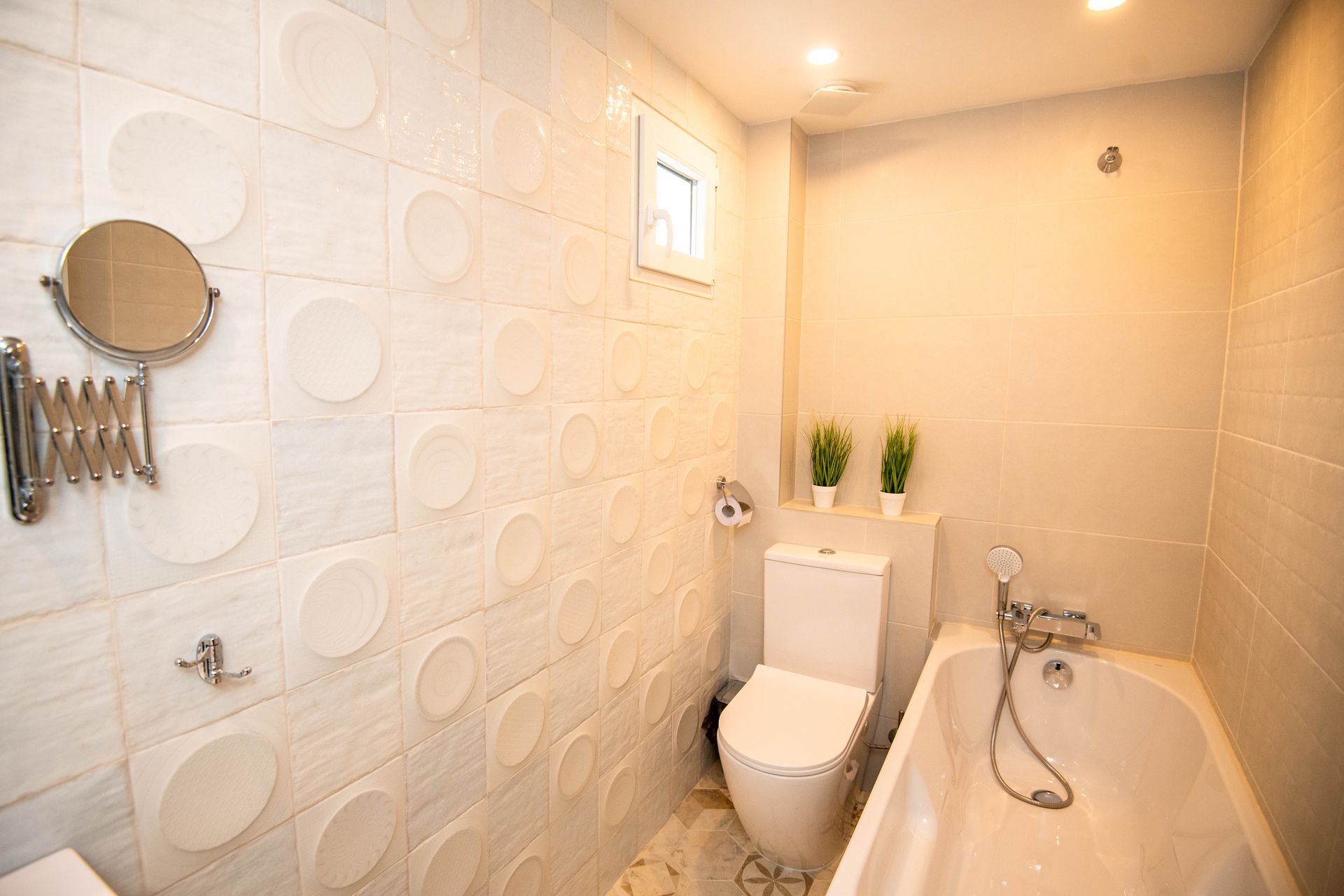 Baño pequeño y blanco con inodoro, bañera y pared de azulejos texturizados.