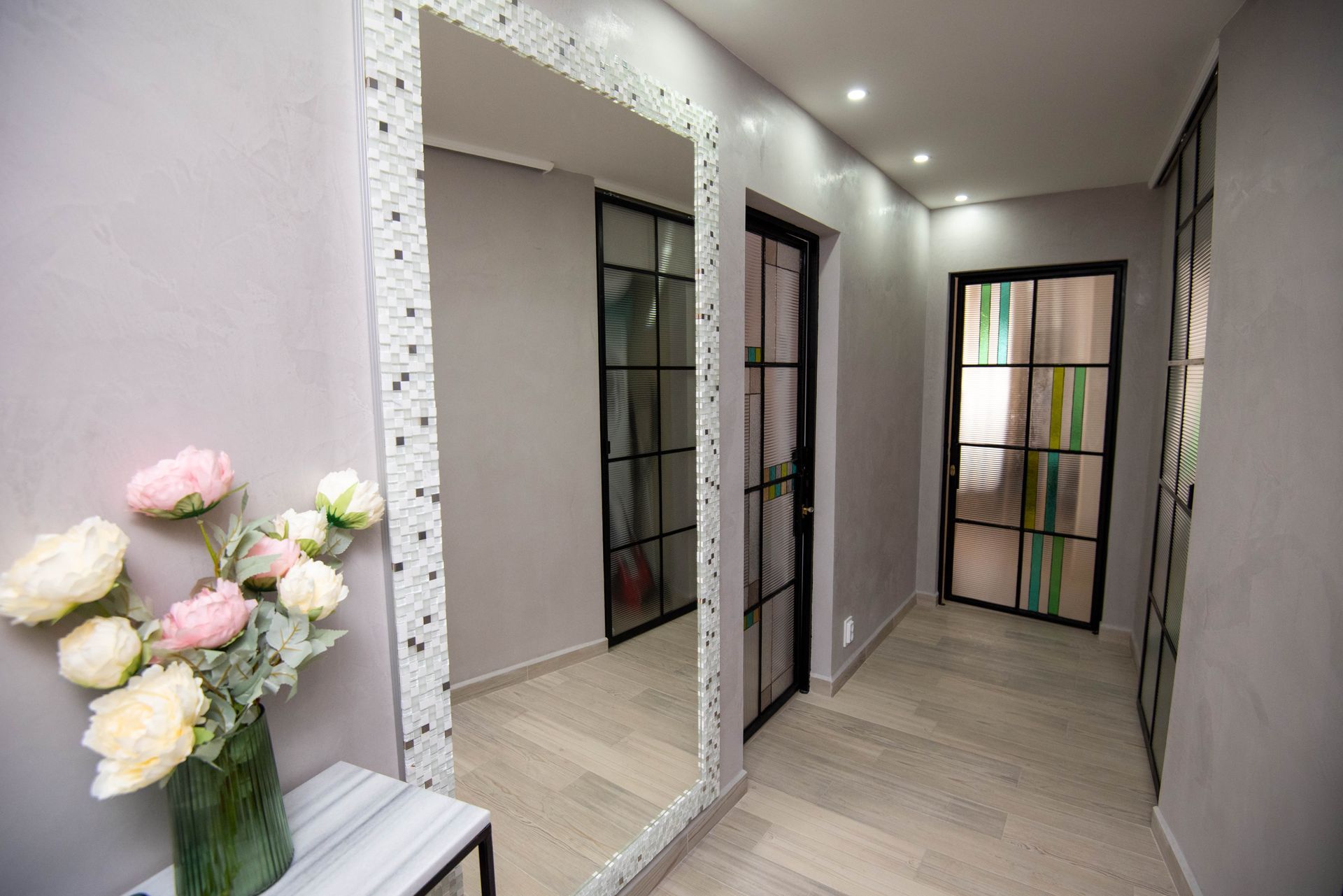 Pasillo con un gran espejo, puertas y un jarrón con flores. Paredes grises y suelo de madera clara.