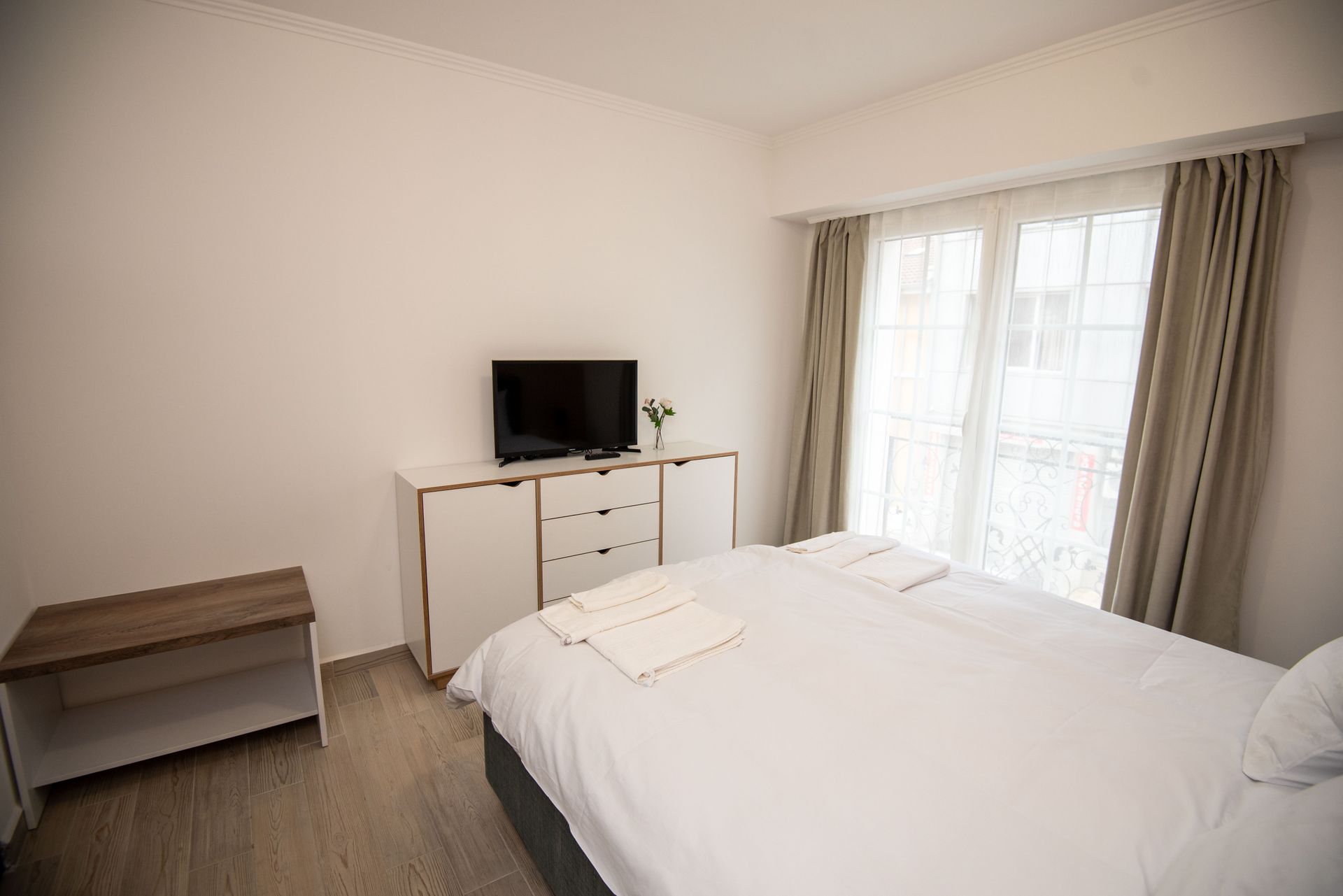 Dormitorio con paredes blancas, cama, cómoda con televisor y ventana con cortinas.