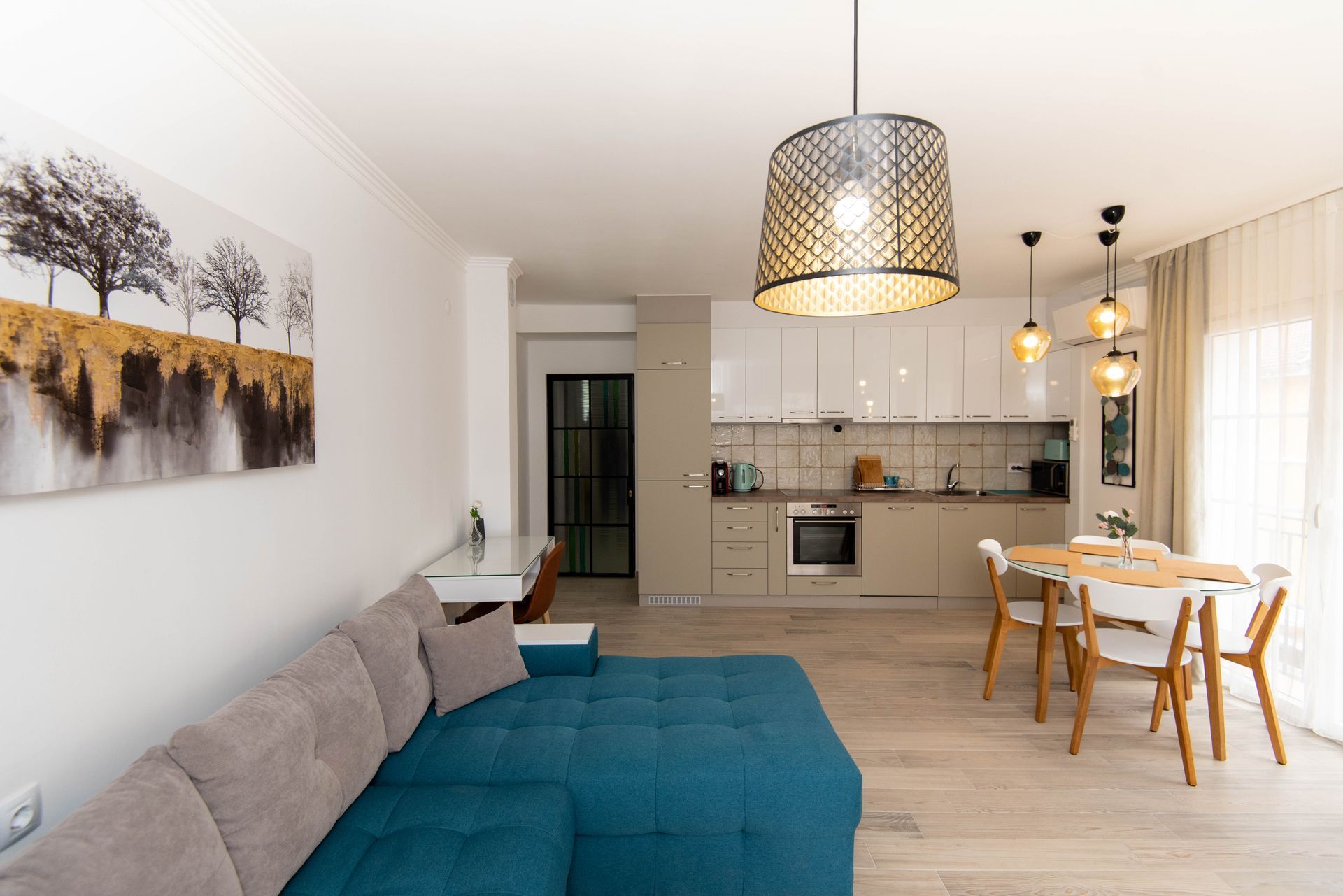 Espacio moderno de planta abierta: sofá color verde azulado, cocina con armarios blancos, mesa de comedor y arte abstracto.