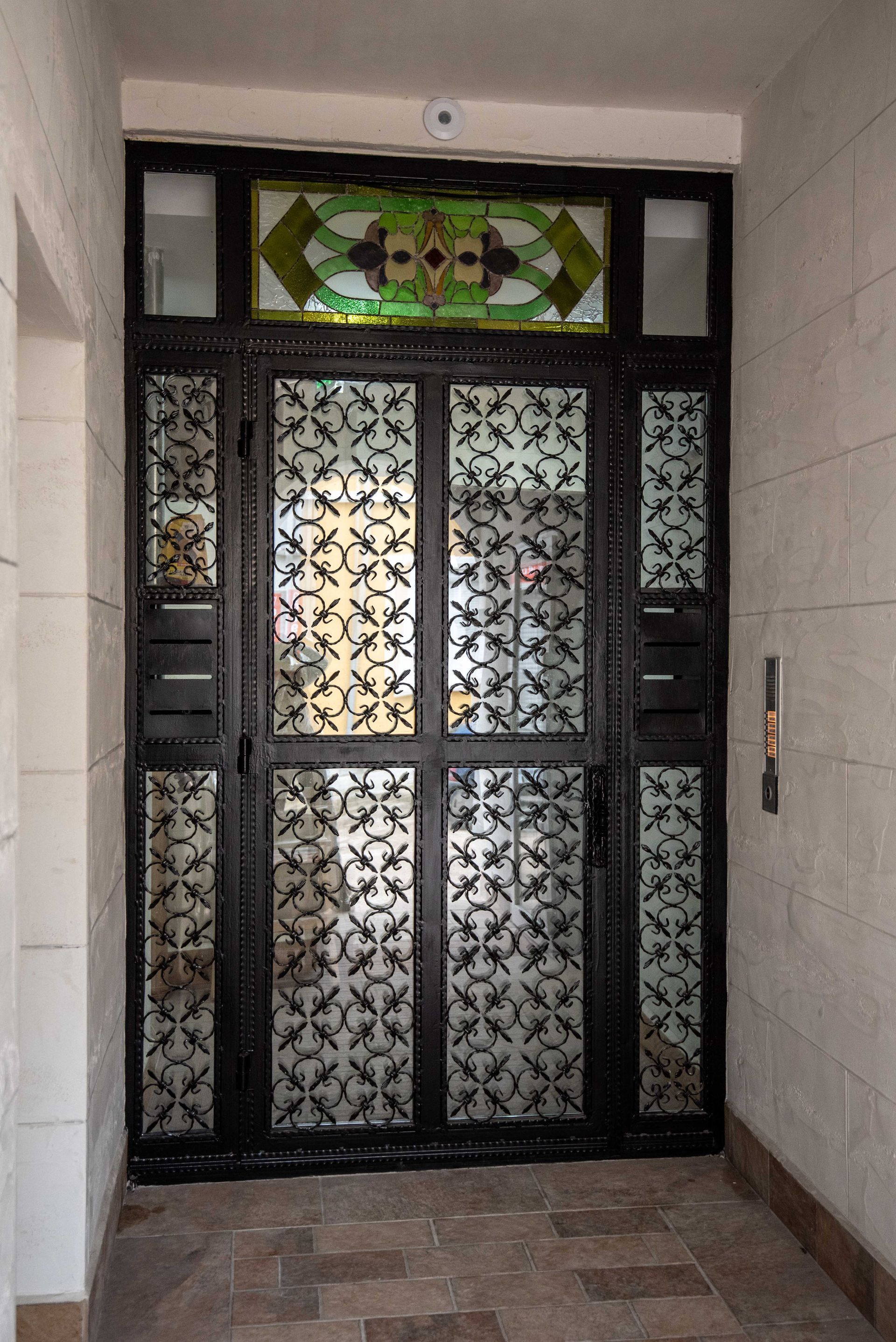 Puerta ornamentada de metal negro con travesaño de vidrieras, situada en una entrada de piedra.