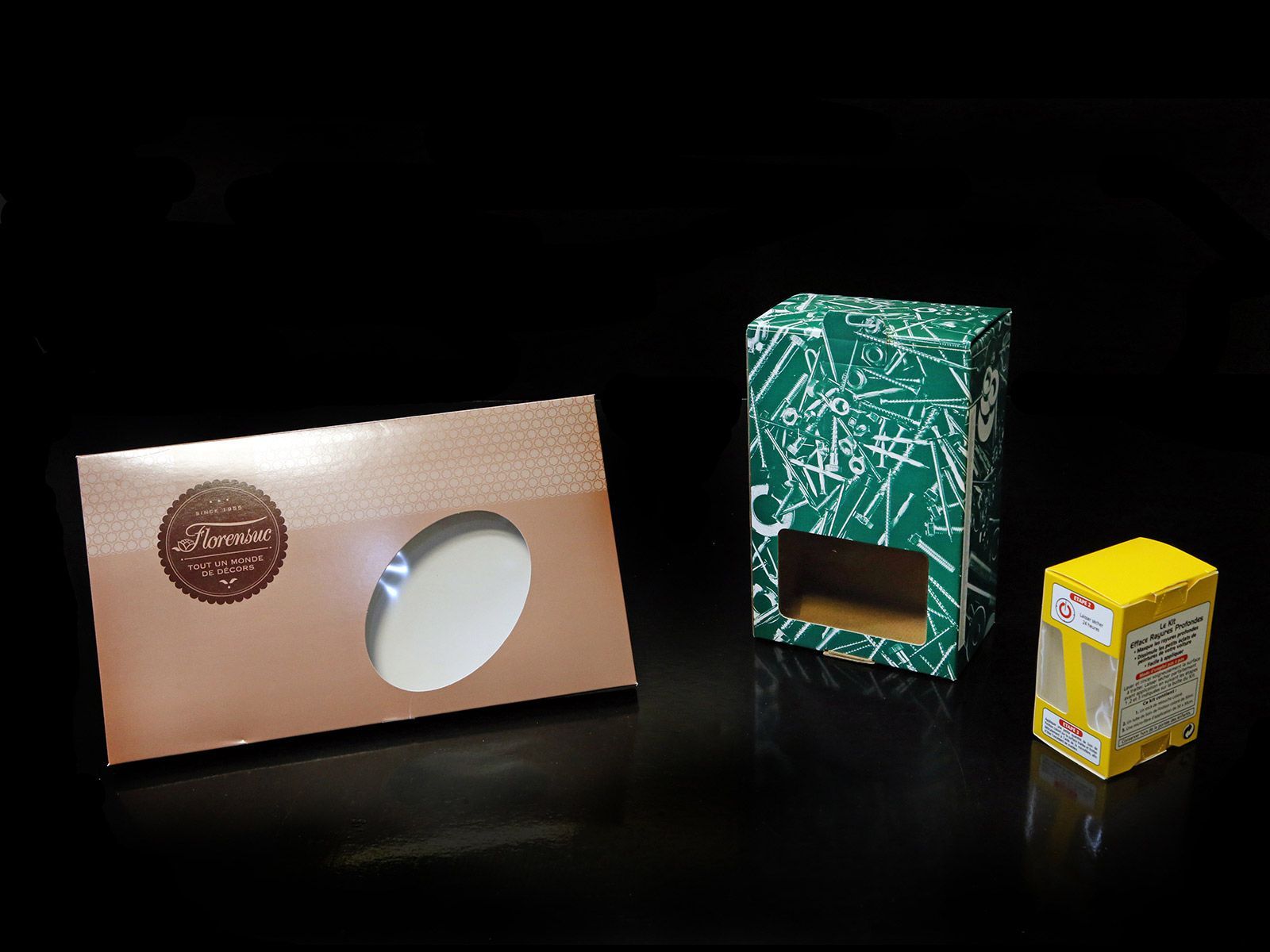 Trois boîtes de produits sur fond noir : bronze, verte à motifs et jaune, chacune avec une fenêtre.