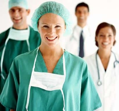 Equipe médica sorridente, vestindo uniformes verdes e jalecos brancos, alguns usando toucas cirúrgicas.