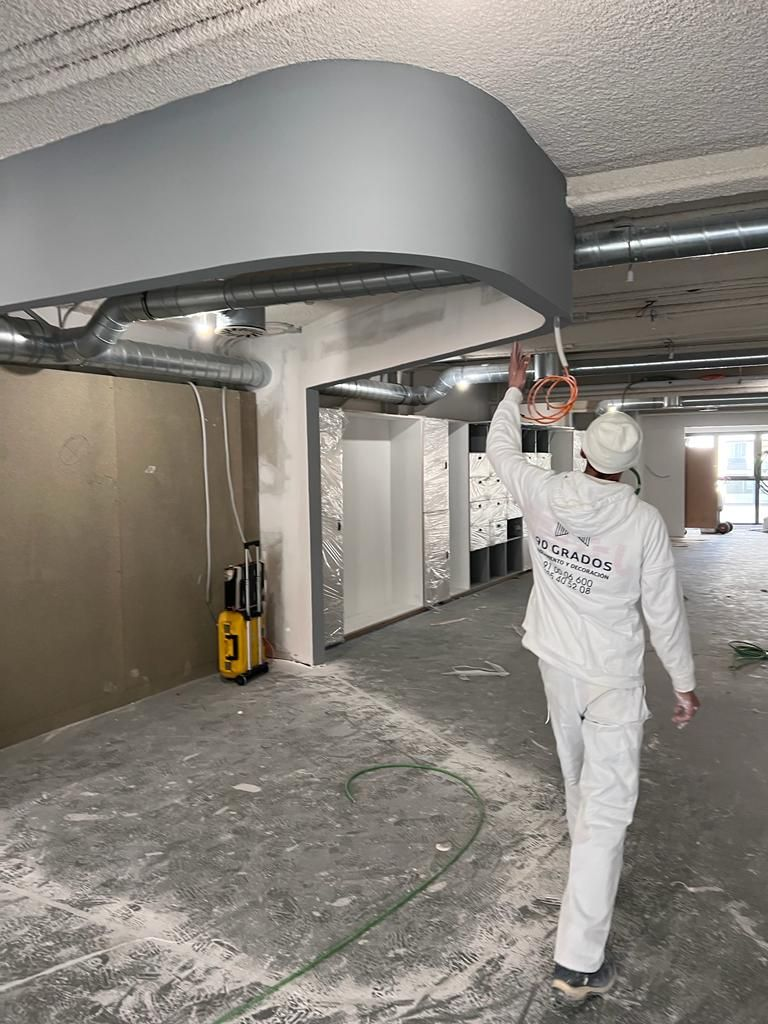 Sitio de construcción interior con infraestructura de techo expuesto, instalación de paneles de yeso y escalera de madera.