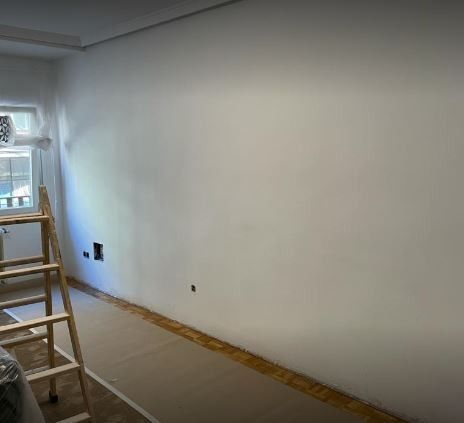 Una habitación vacía pintada de blanco, con una escalera de madera, enchufes eléctricos y suelo encintado.