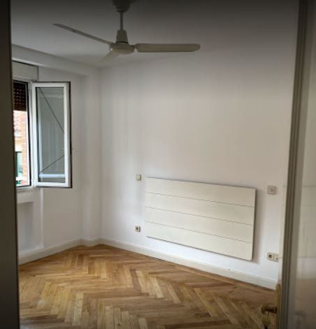 Habitación vacía con suelo de parqué, paredes blancas y una ventana con persianas. Se ven un ventilador de techo y una estufa de pared.