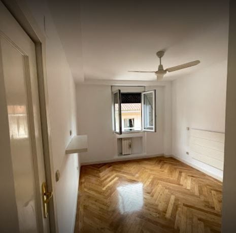 Habitación vacía con suelo de parqué, paredes blancas y una ventana con vistas despejadas. Dispone de ventilador de techo y radiador.