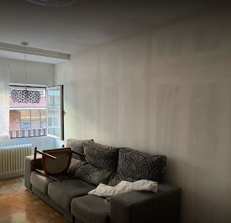 Una sala de estar con un sofá gris, una ventana y una silla. Las paredes son de color claro y se ve un balcón por la ventana.