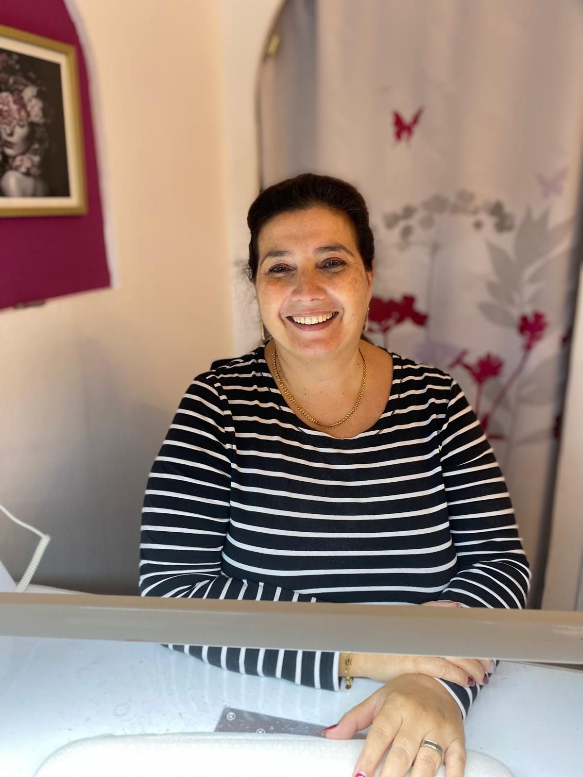 Eine Frau in einem gestreiften Hemd lächelt an einem Schreibtisch. Im Hintergrund sind eine rosa Wand, Kunstwerke und ein Vorhang zu sehen.